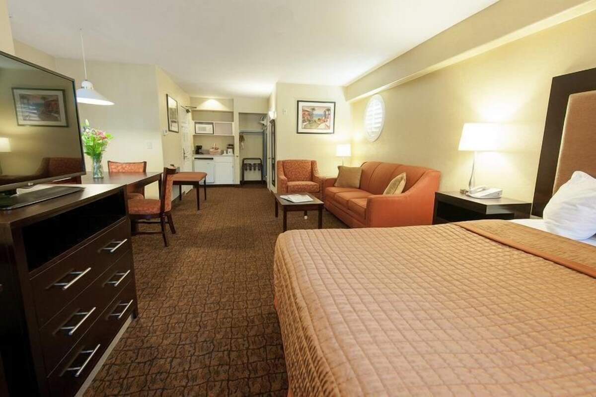 A hotel room with a bed, a desk, a chair, a sofa, and a kitchenette in the background.