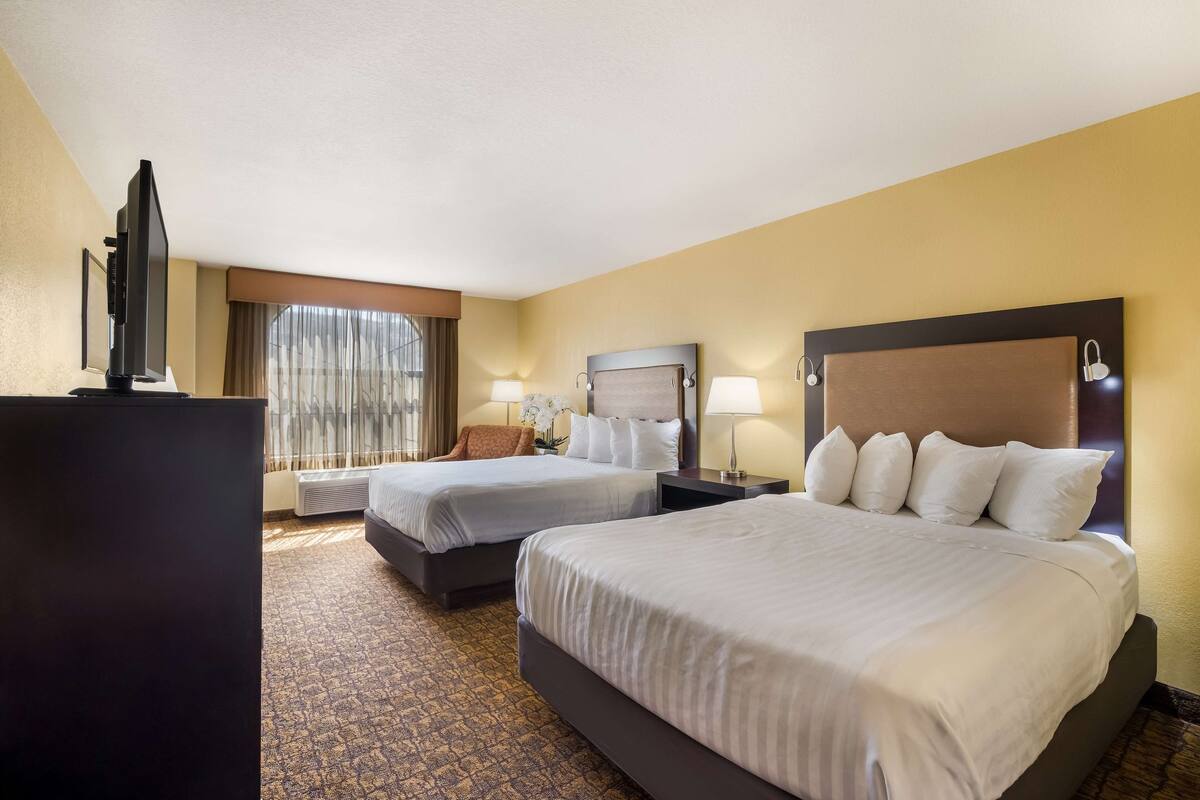 A hotel room with two beds, a flat-screen TV, a desk, and a large window with curtains.