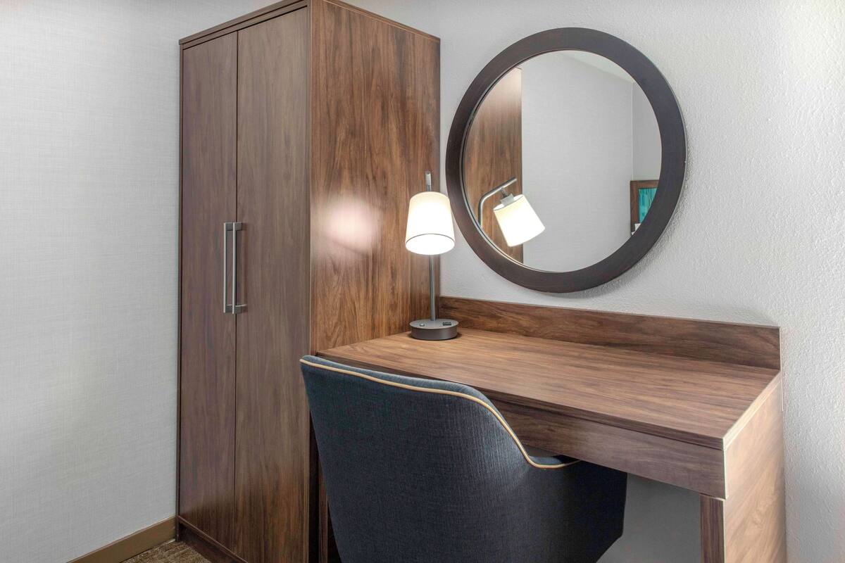 A wooden desk with a round mirror and a lamp, a dark upholstered chair, and a wooden wardrobe.