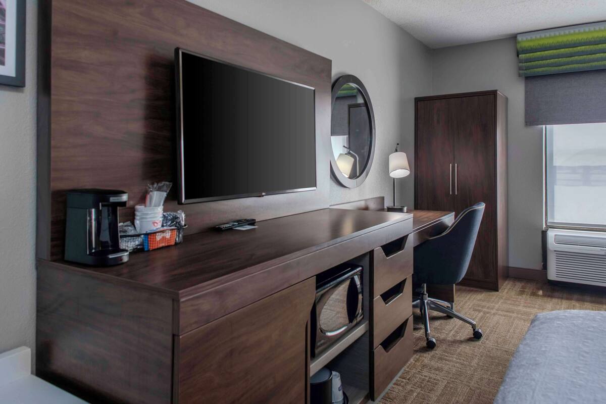 A hotel room with a wooden desk, a flat-screen TV, a mirror, a lamp, a wardrobe, and a bed.