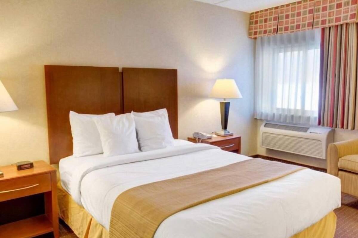 Hotellihuone, jossa on suuri sänky, yöpöytä, lamppu, tuoli ja ikkuna, jossa on verhot.