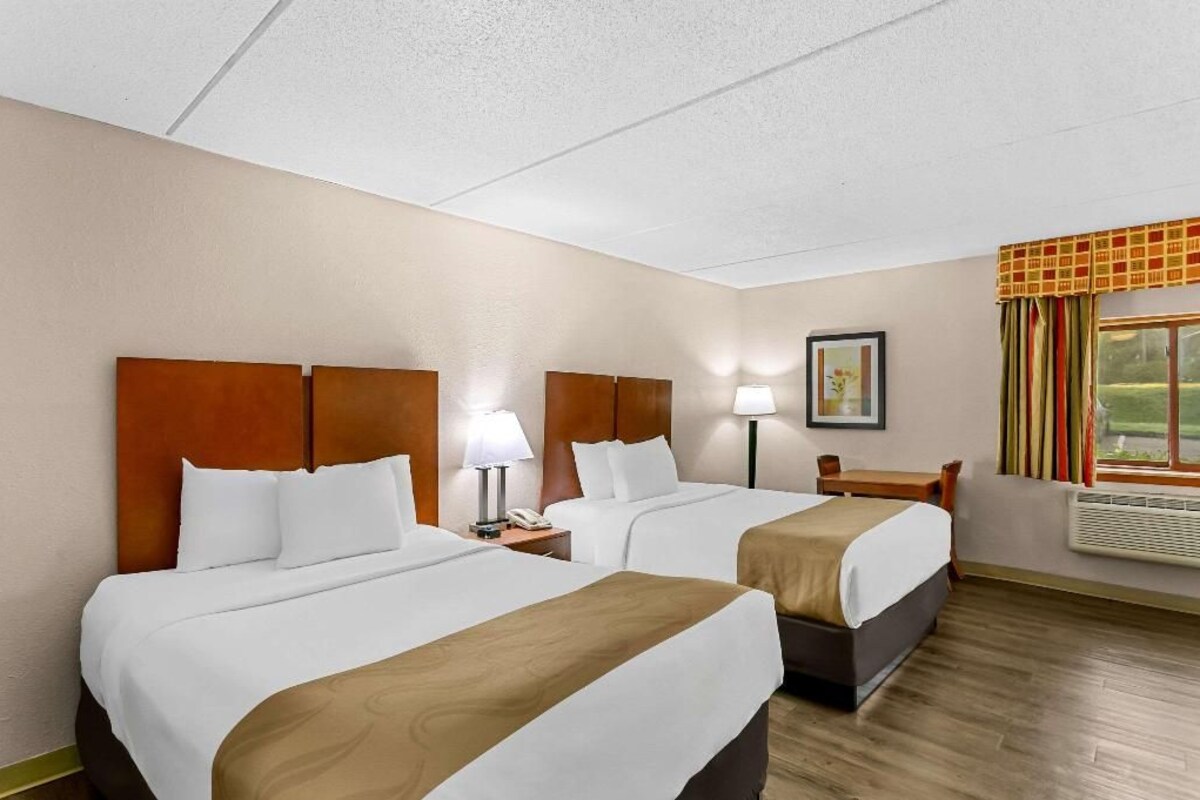 A hotel room with two beds, a desk, a chair, a window with curtains, and a framed picture on the wall.