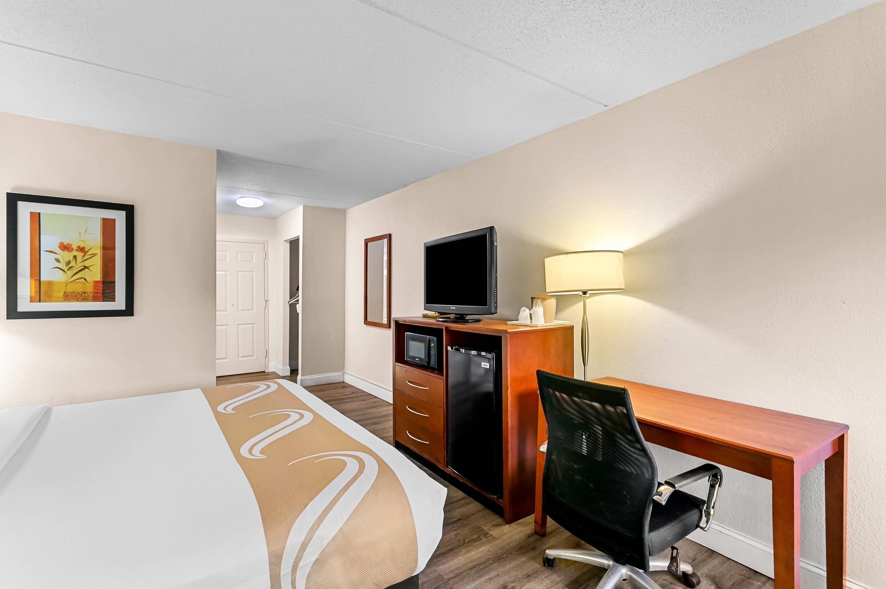 A hotel room with a bed, a desk with a computer, a television, a refrigerator, and a lamp.