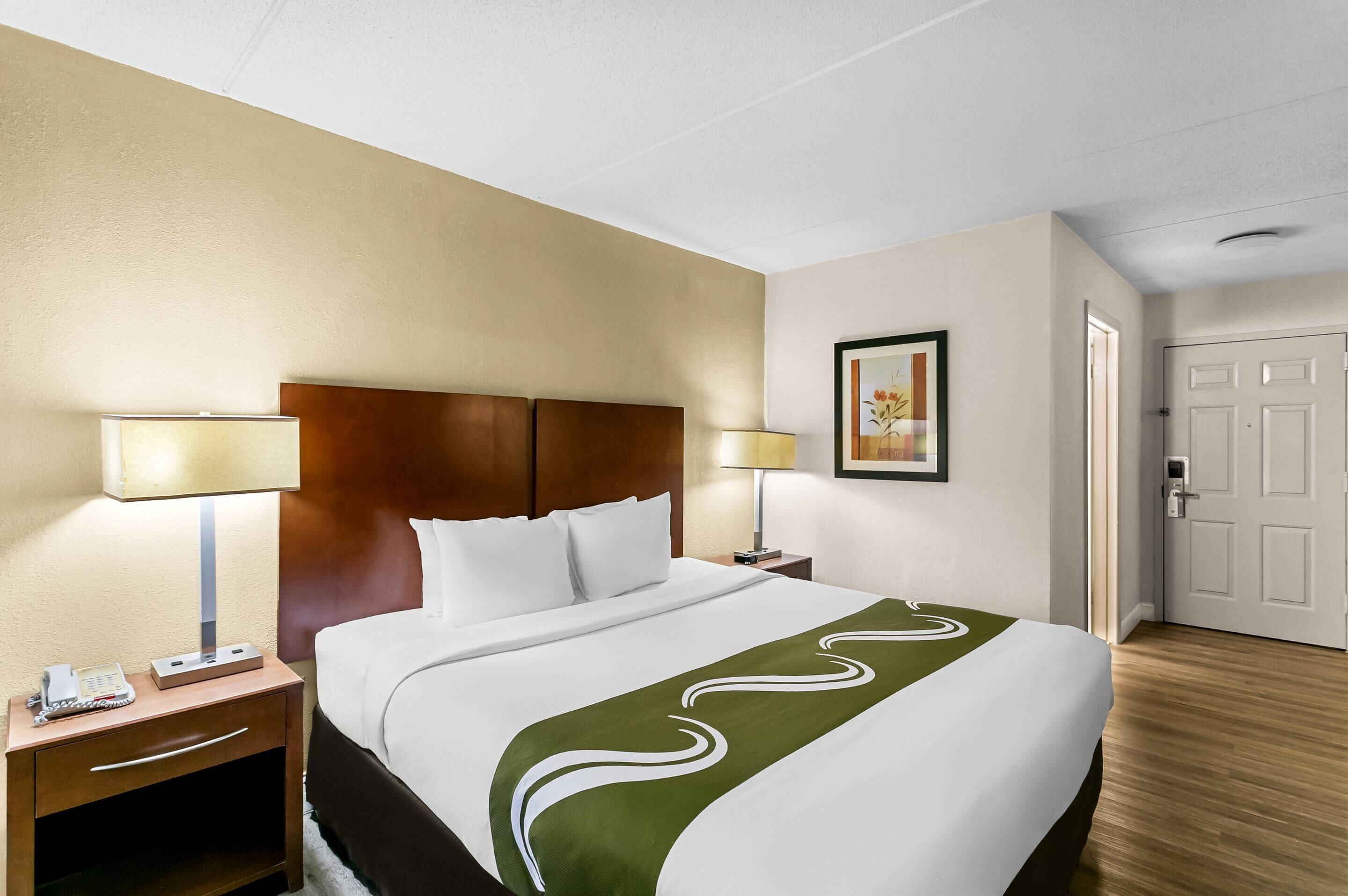 Habitación de hotel con cama, lámparas de noche, mesita de noche, un cuadro en la pared y una puerta.