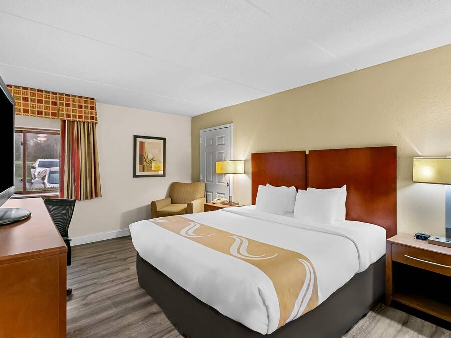 A hotel room with a bed, a desk with a television, a chair, a nightstand with a lamp, and a door.