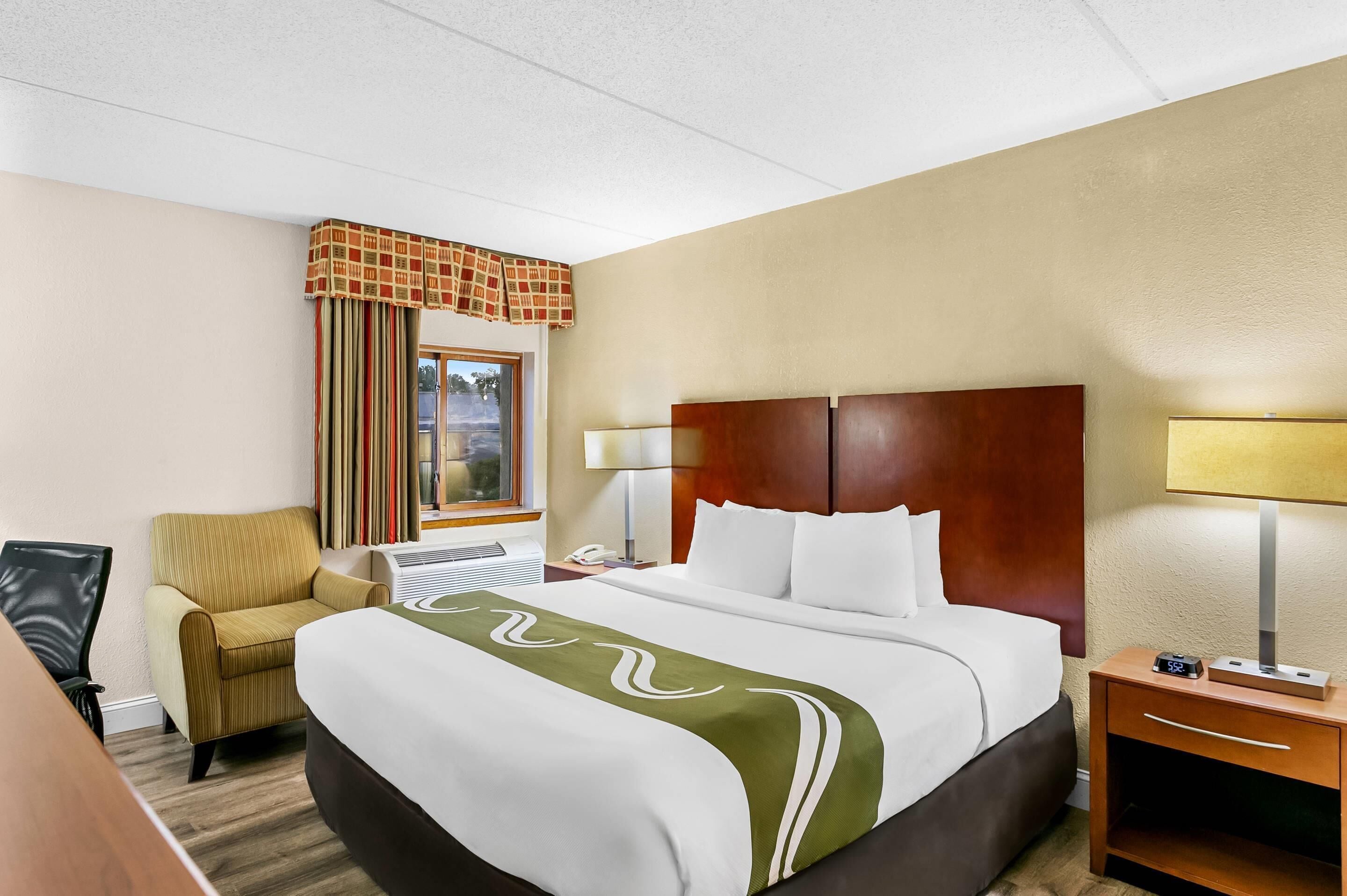 A hotel room with a bed, a chair, a desk, a lamp, and a nightstand.