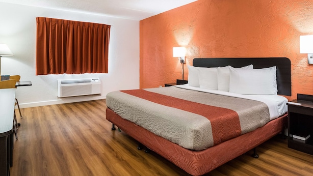 A hotel room with a large bed, an air conditioning unit, a desk with a chair, and a window with curtains.