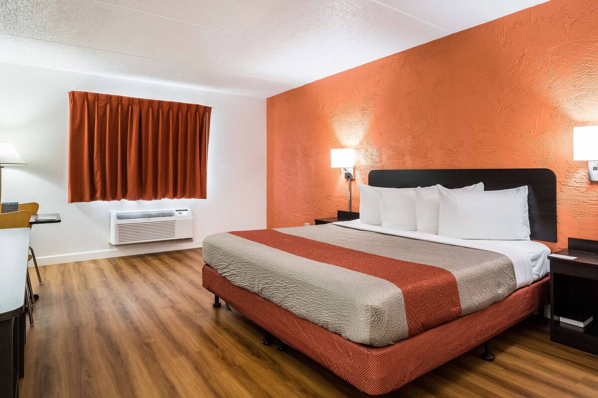 A hotel room with a large bed, an air conditioning unit, a desk with a chair, and a window with curtains.