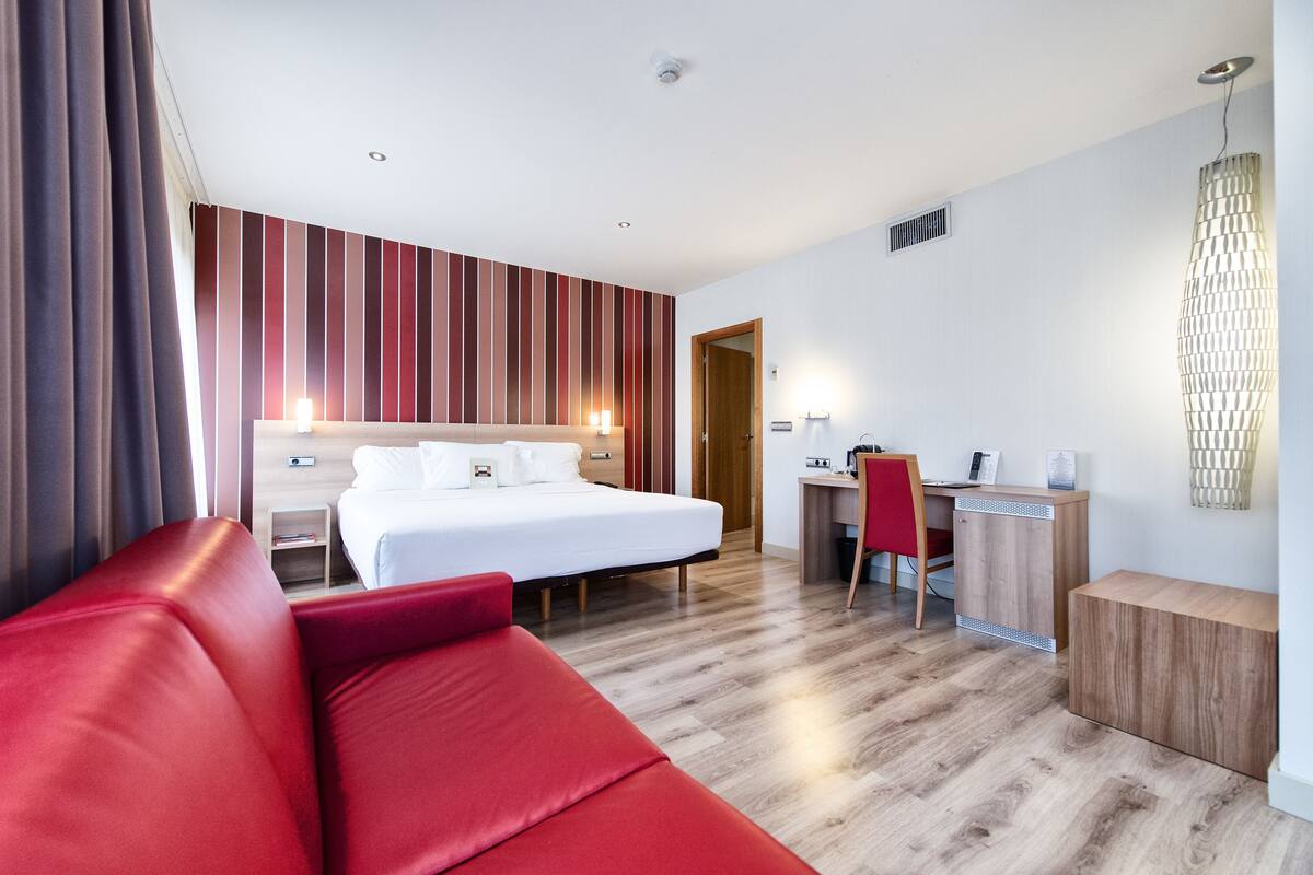A hotel room with a bed, a desk, a red sofa, and a wooden cabinet.