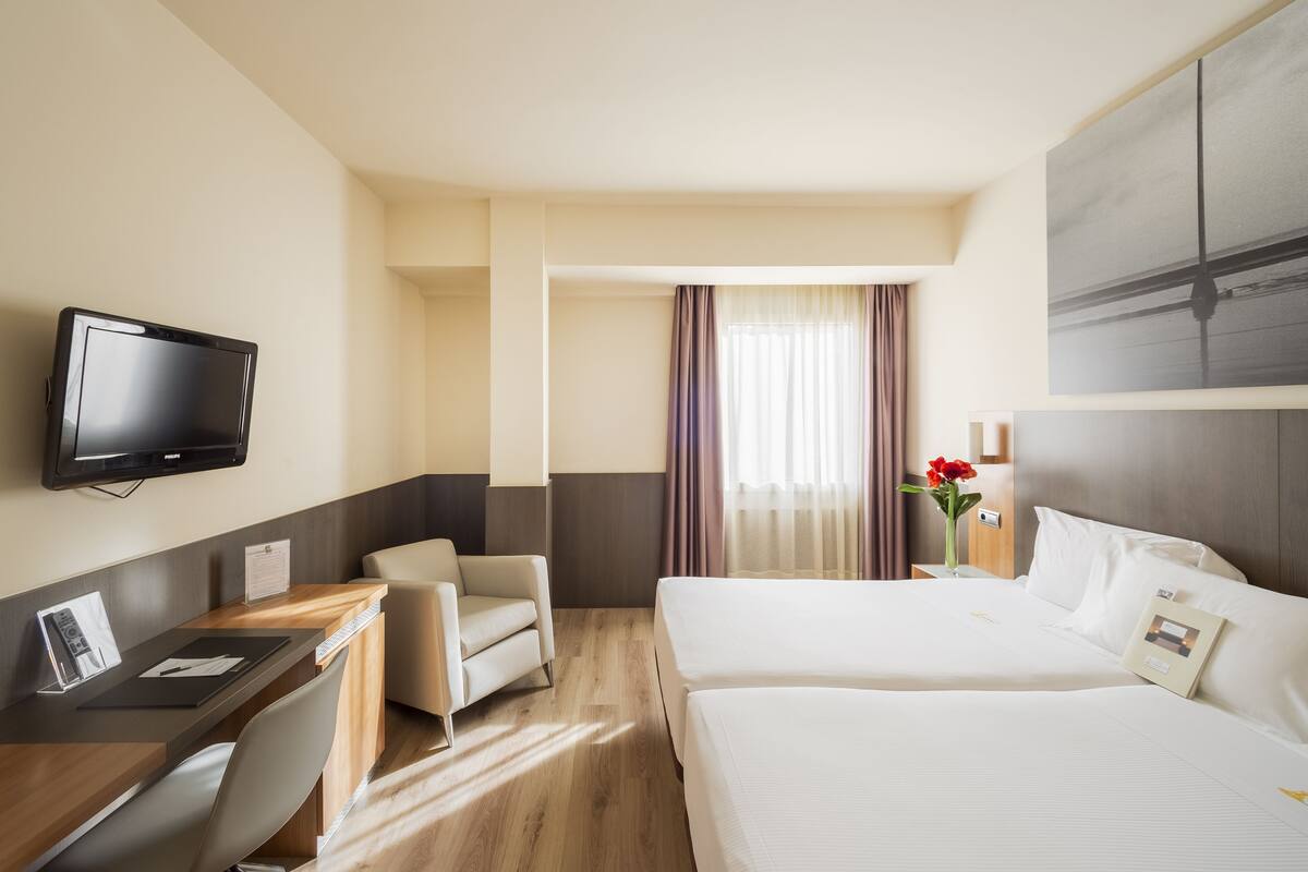 A hotel room with a desk, two beds, a TV, and a view of the outside.