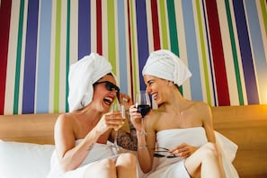 Two people sitting on a bed with striped wallpaper, holding wine glasses.
