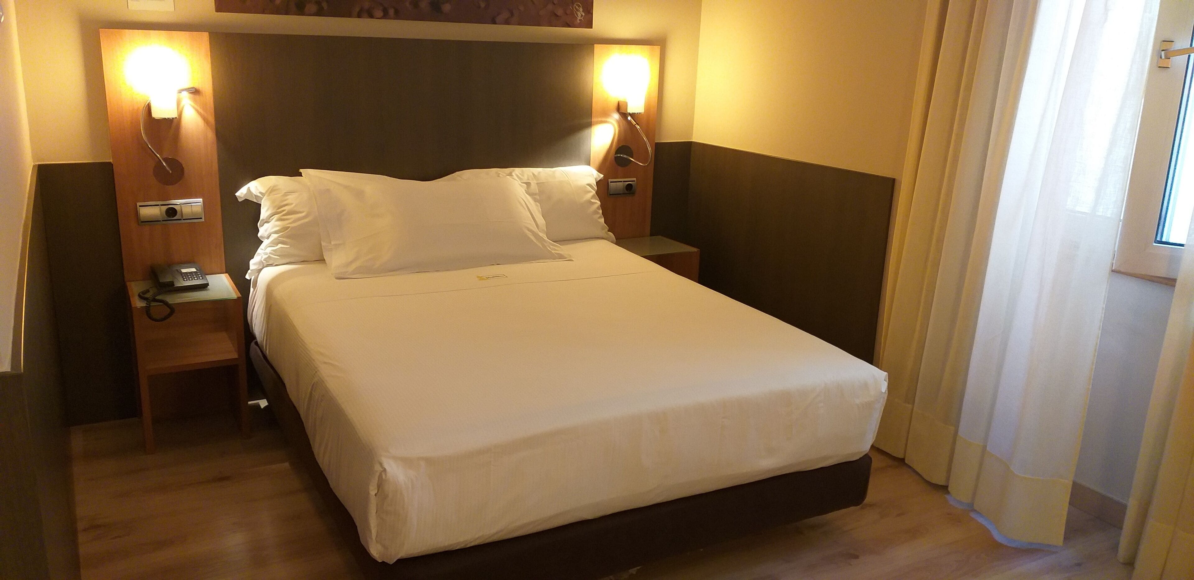 A hotel room with a bed, two bedside lamps, a bedside telephone, and a window with curtains.