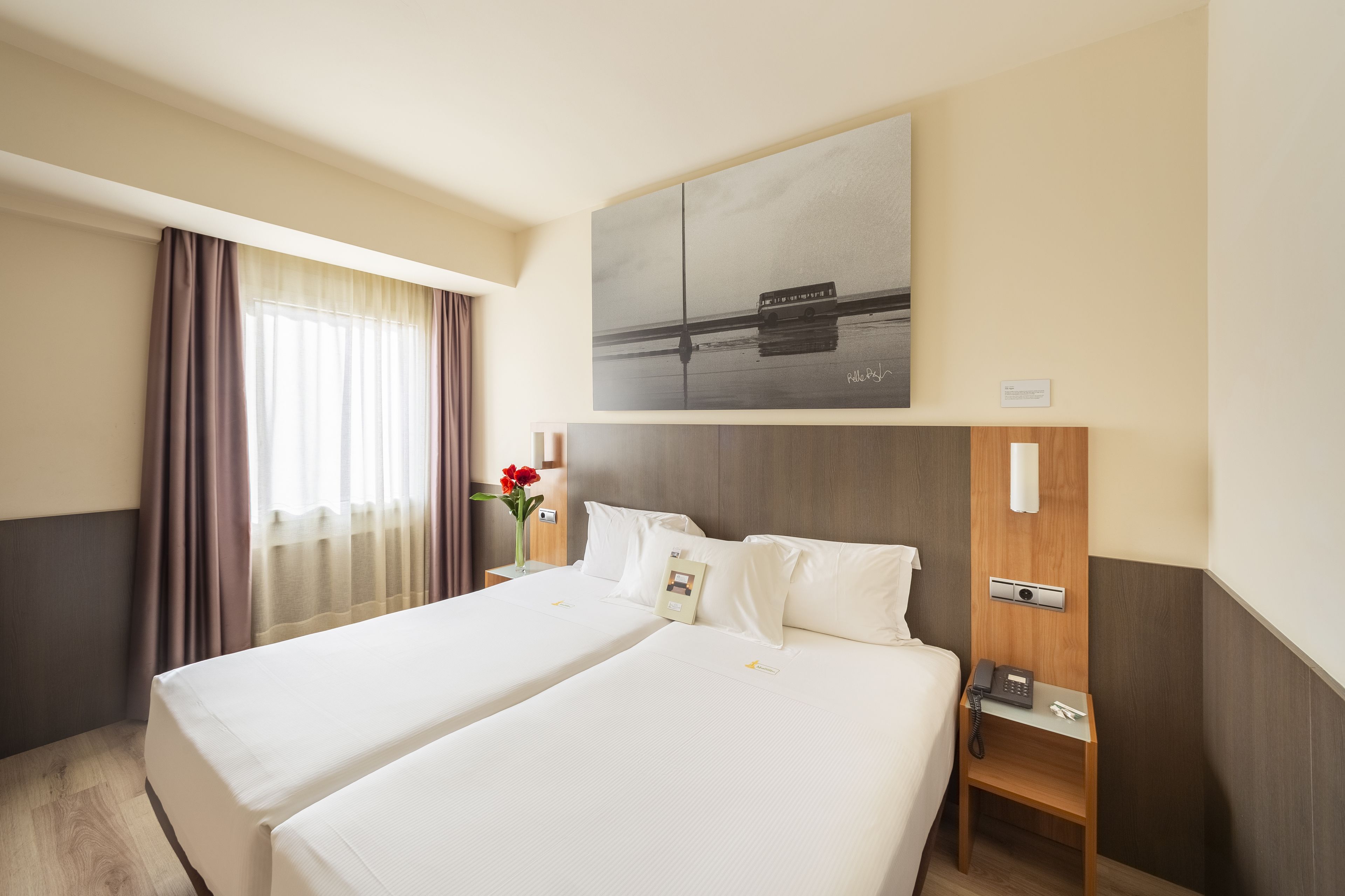 A hotel room with two beds, a wooden nightstand with a phone, a painting above the beds, and a window with curtains.