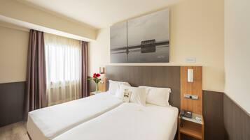 A hotel room with two beds, a wooden nightstand with a phone, a painting above the beds, and a window with curtains.