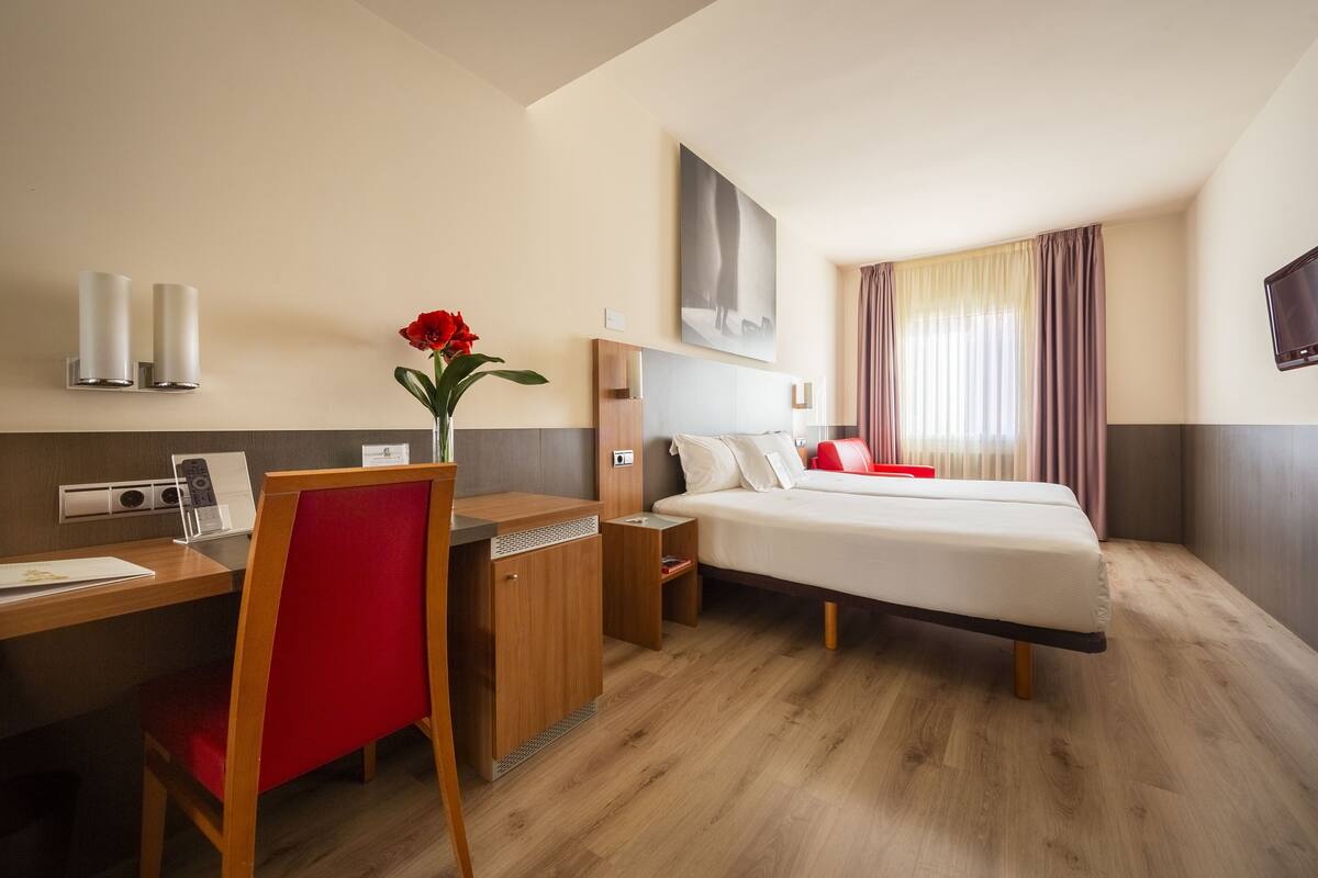 A hotel room with a bed, desk, chair, bedside table, and a vase with a flower.