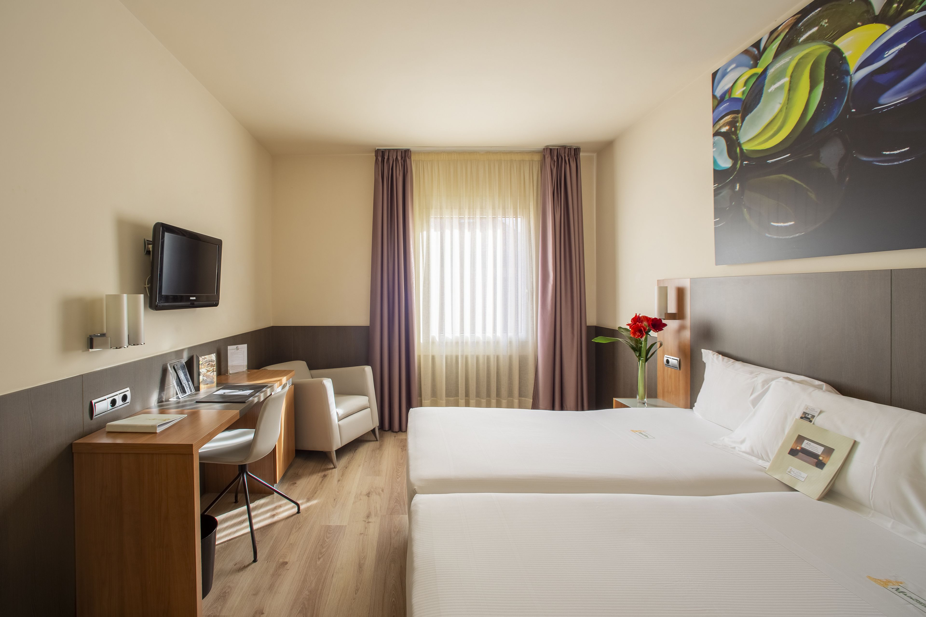 A hotel room with a bed, a desk with a chair, a television mounted on the wall, and a window with curtains.