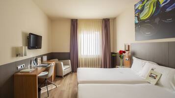 A hotel room with a bed, a desk with a chair, a television mounted on the wall, and a window with curtains.