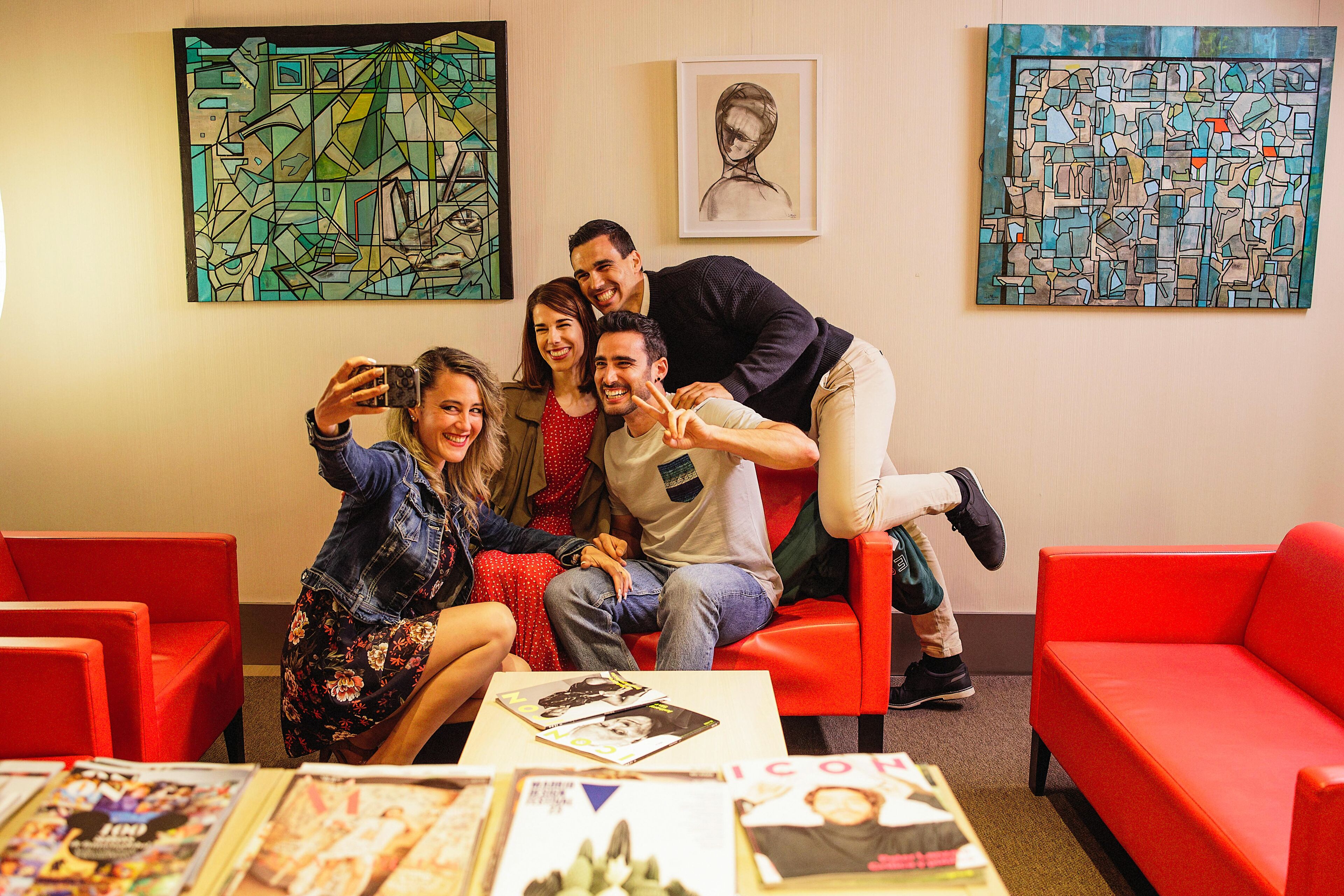 Un gruppo di persone sedute su un divano rosso in una stanza con dipinti astratti alle pareti e un tavolino con riviste in primo piano.