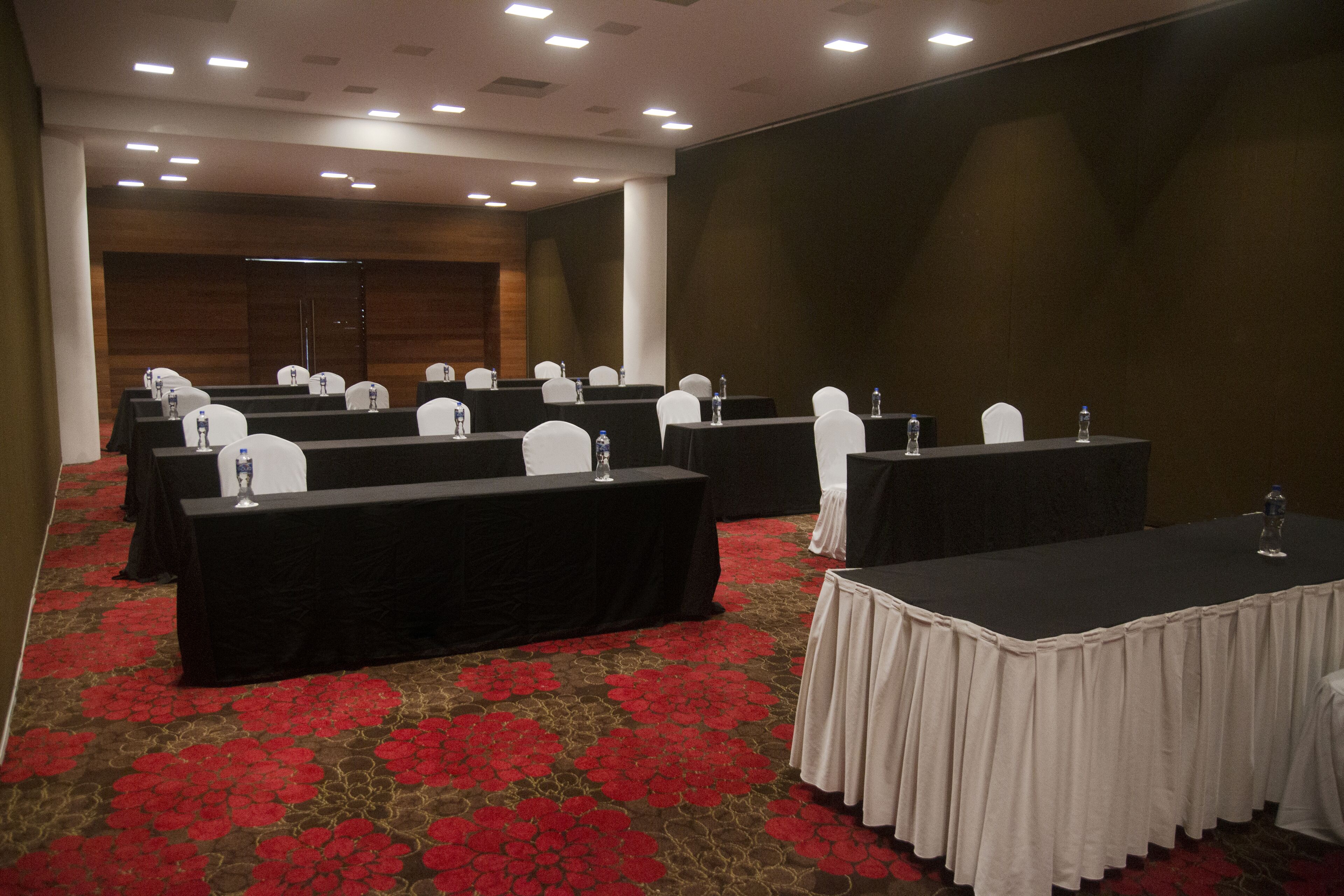 Une salle de conférence dotée de plusieurs tables, chacune recouverte d’une nappe noire et entourée de chaises blanches. La salle est agrémentée d’un tapis rouge à motifs floraux et est équipée de luminaires encastrés au plafond.