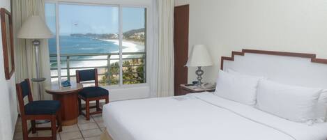 Un cuarto de hotel con una cama grande, una mesita con sillas, una ventana con vista al océano y una mesita de noche con lámpara.