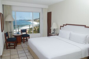 Un cuarto de hotel con una cama grande, una mesita con sillas, una ventana con vista al océano y una mesita de noche con lámpara.
