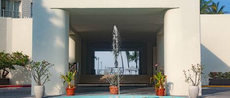 Zona de piscina con una fuente, plantas en macetas y vistas a un edificio al fondo.