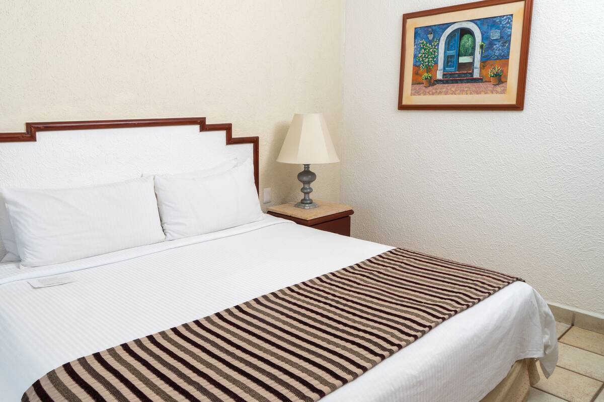 Una cama bien hecha con ropa de cama blanca y una manta de rayas, una lámpara de noche y un cuadro en la pared.