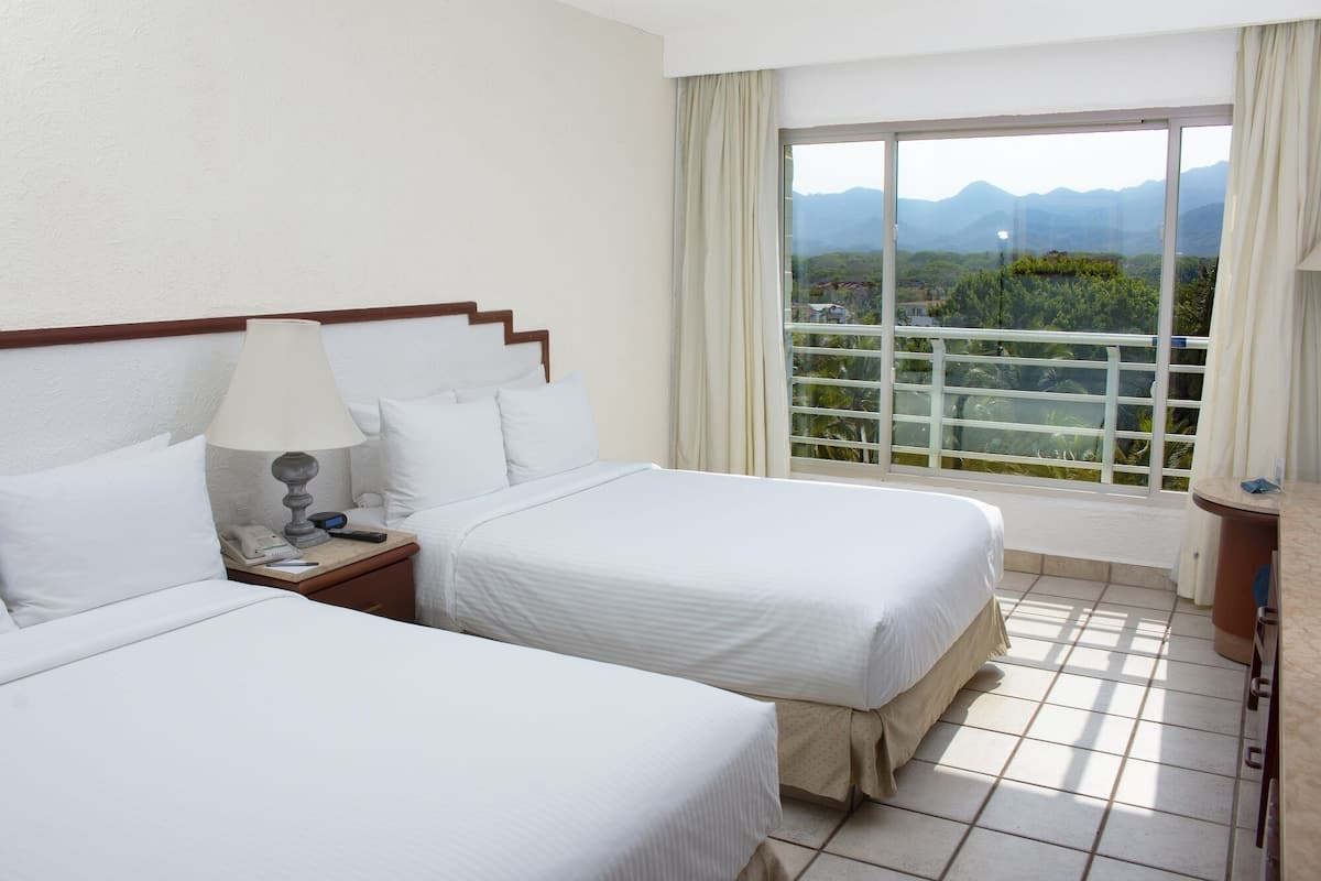 Habitación de hotel con dos camas, una mesita de noche con lámpara, una ventana con vistas a montañas y un balcón con barandilla.