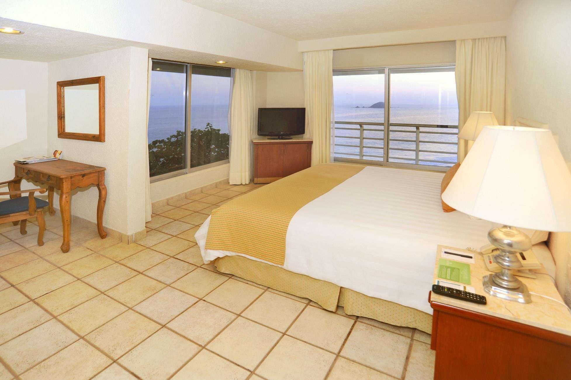 A hotel room with a bed, a TV, a desk, and a view of the sea.