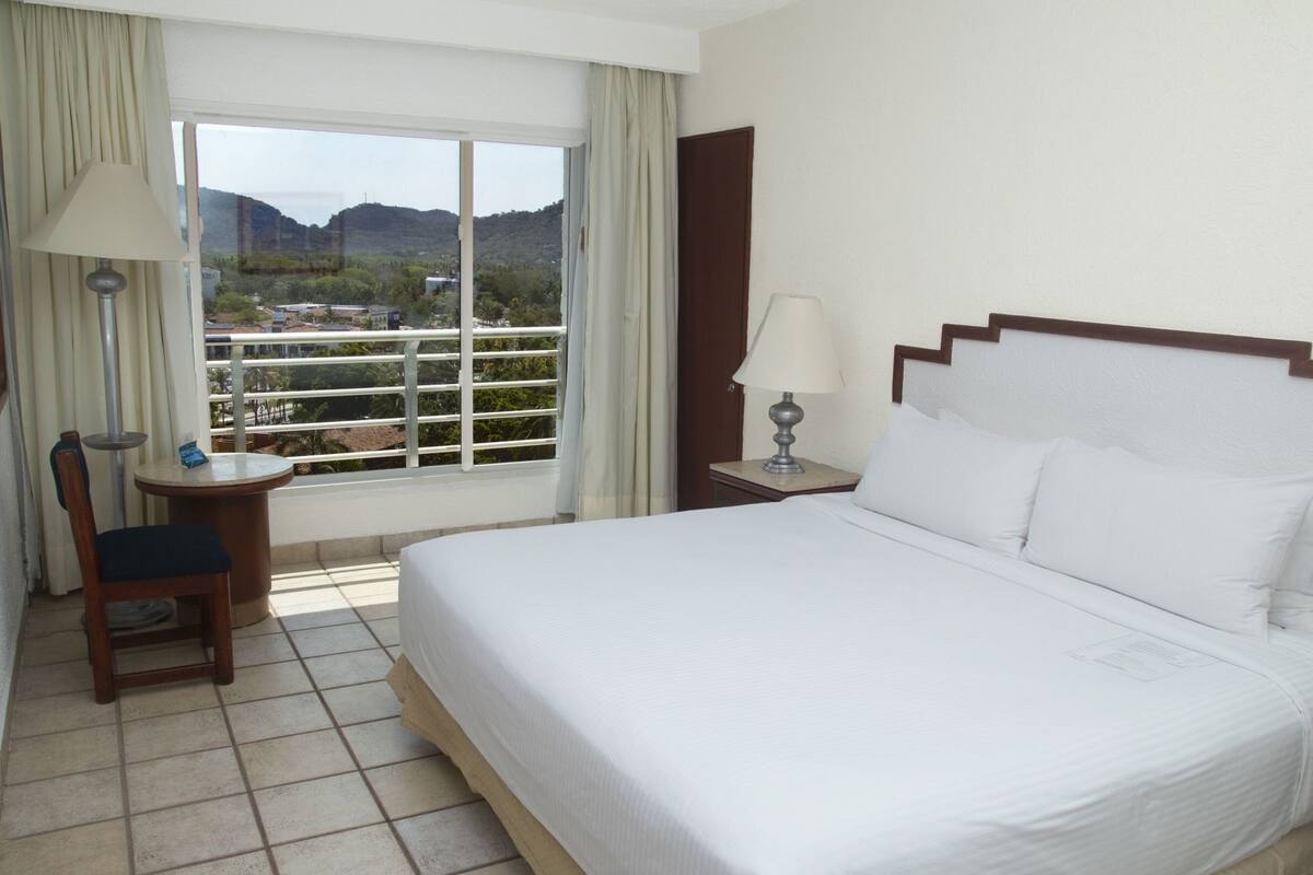 Un cuarto de hotel con una cama, una silla, una mesa, una lámpara y un ventanal con vistas al paisaje.