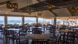 Un comedor espacioso con mesas y sillas múltiples, ventanas amplias que ofrecen vista al océano y la playa, y lámparas colgantes de mimbre.