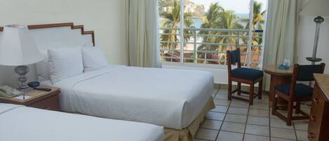 A hotel room with two beds, a desk, a chair, a lamp, and a view of the beach and palm trees.