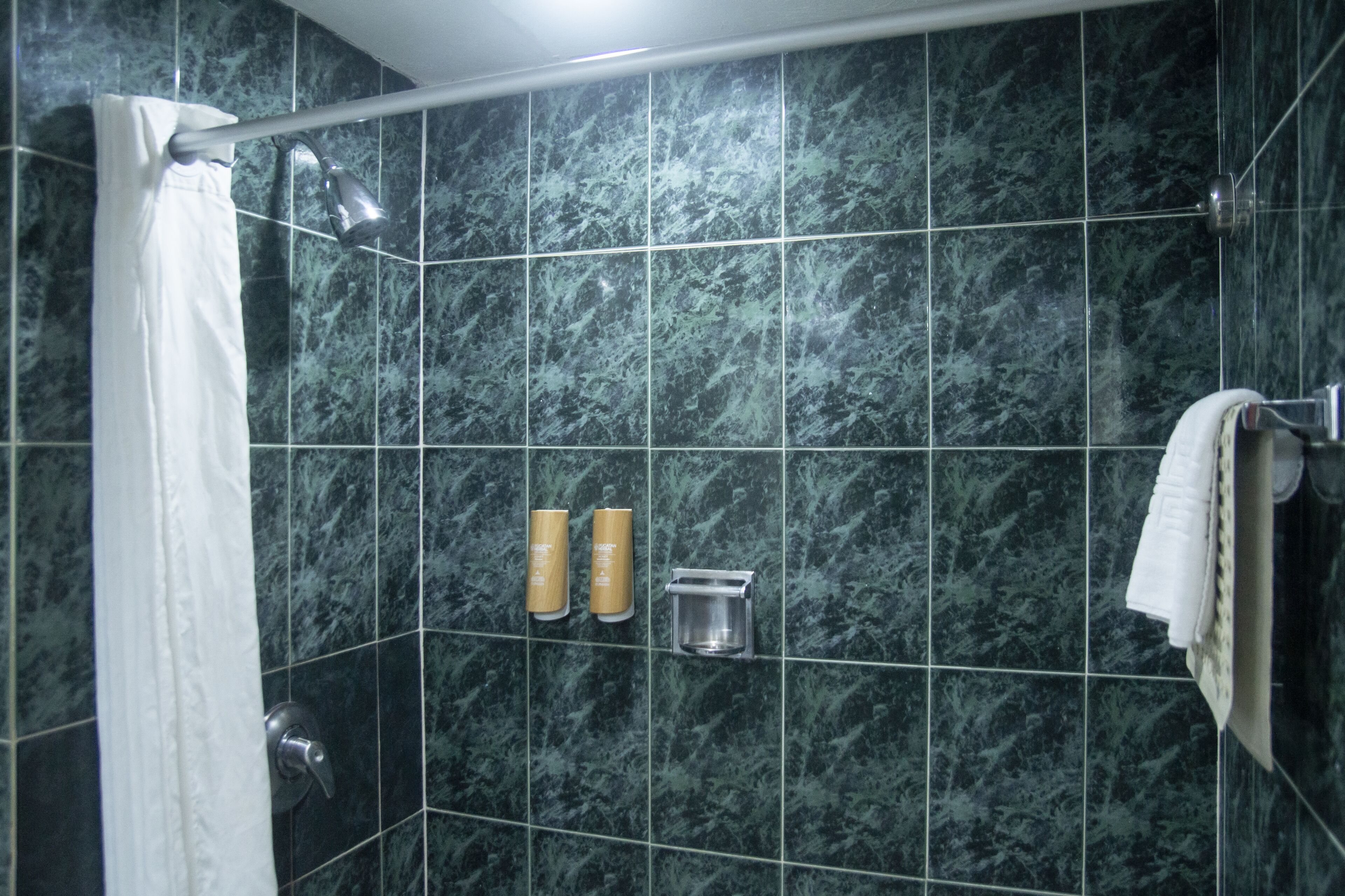 Un baño con baldosas de mármol verde oscuro, una cortina de ducha, un perchero y dos botellas de champú y acondicionador.