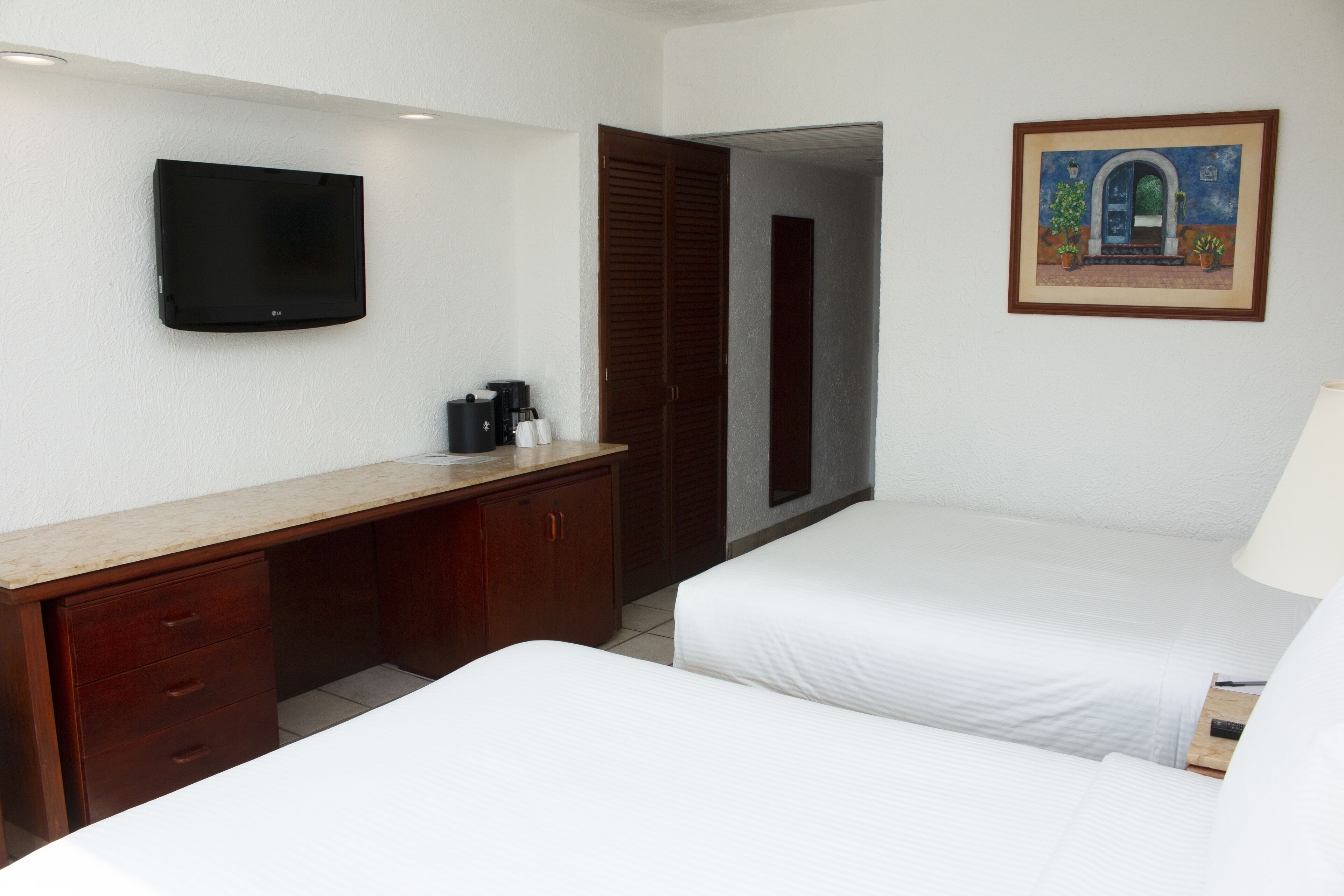 Habitación de hotel con televisor de pantalla plana, un escritorio de madera con cajones, cafetera, dos camas y un cuadro en la pared.