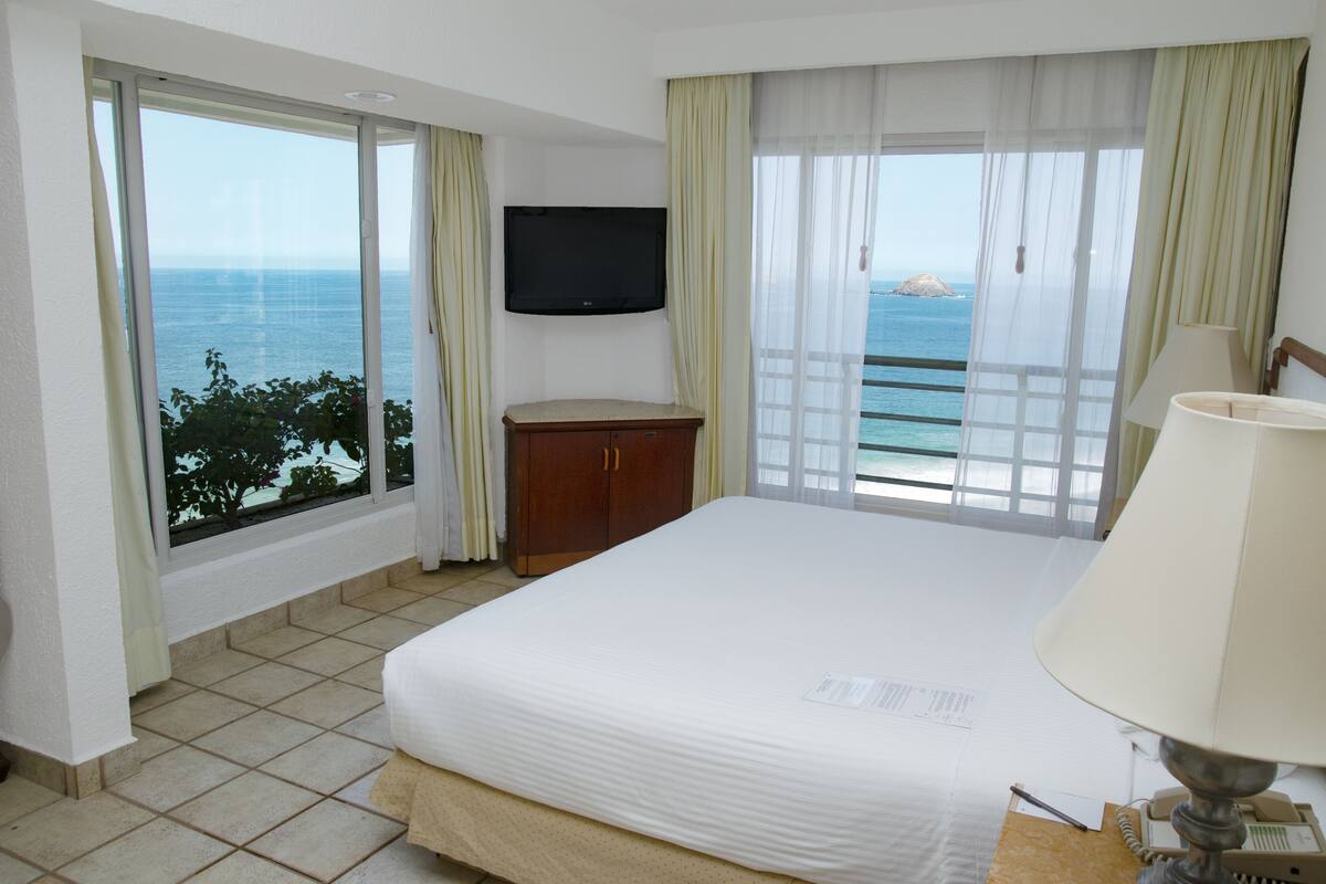 Habitación de hotel con cama, televisor, aparador de madera, lámpara y vista al océano.