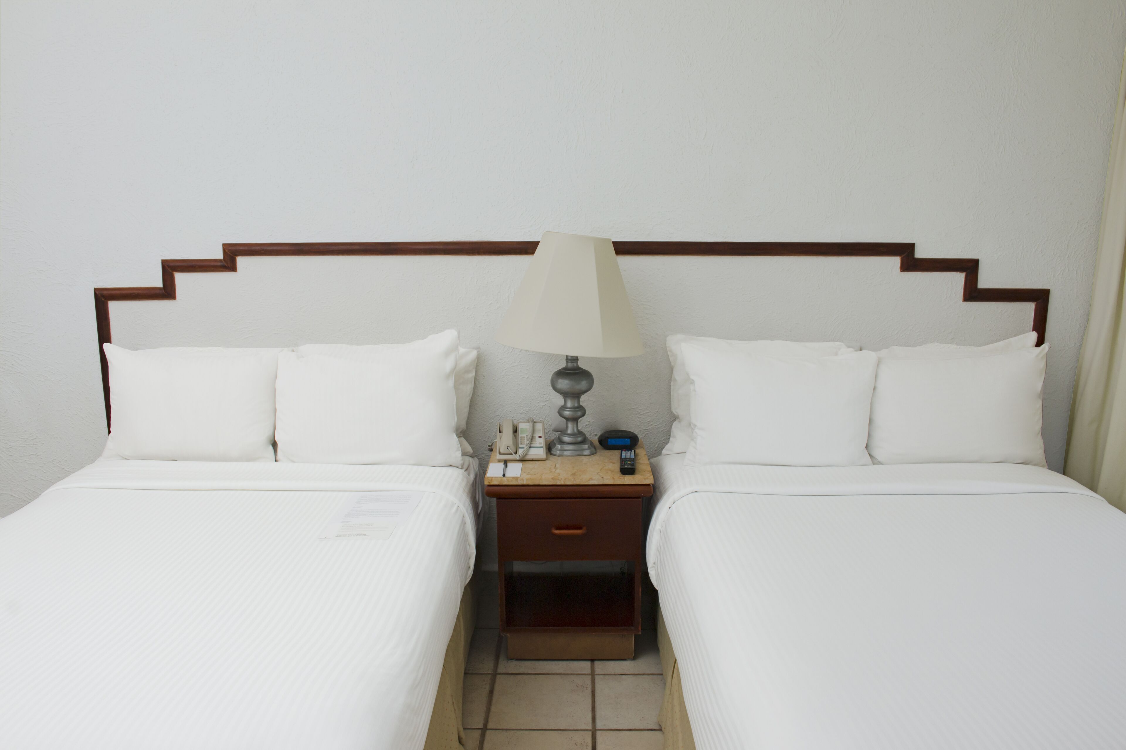 A double bed with white linens, a wooden nightstand with a lamp, and a small drawer.