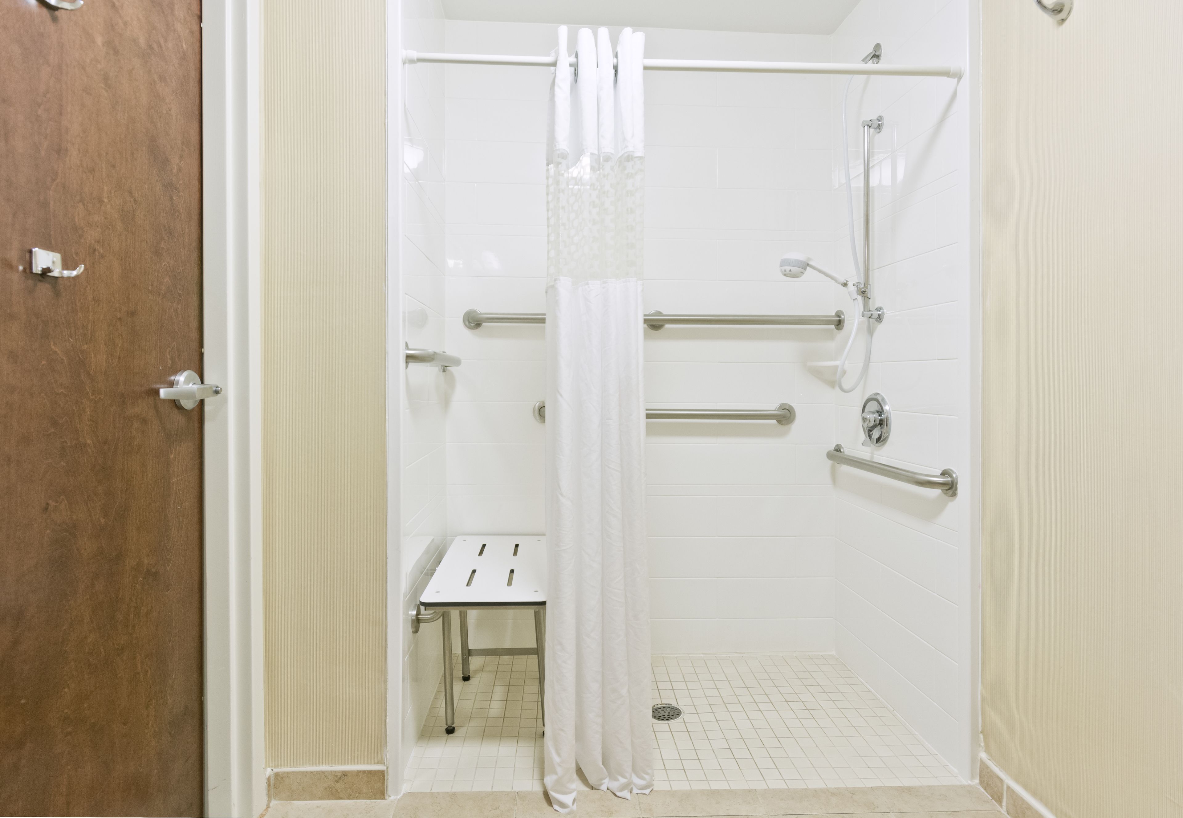Un baño con ducha, barras de apoyo, suelo de azulejos blancos y una puerta.