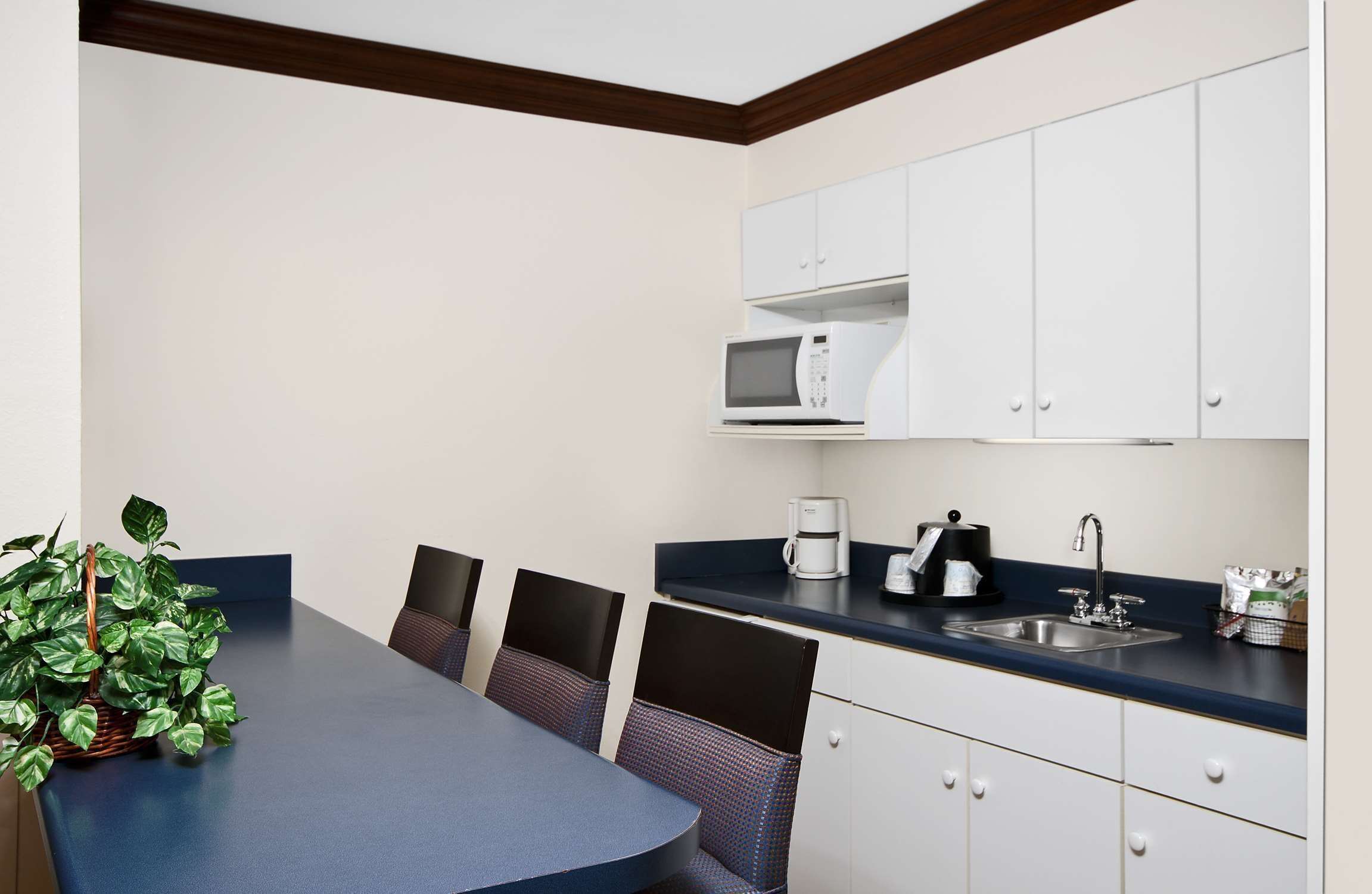 A compact kitchen with white cabinets, a microwave, a coffee maker, and a sink. There is a table with chairs and a potted plant.