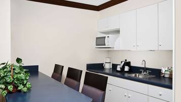 A compact kitchen with white cabinets, a microwave, a coffee maker, and a sink. There is a table with chairs and a potted plant.