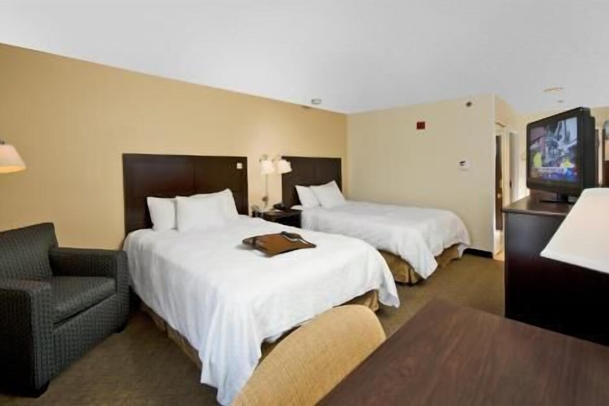A hotel room with two beds, a desk, a chair, a TV, and a lamp.