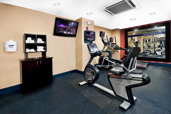 Un gimnasio con cintas de correr, bicicletas estáticas y un espejo grande. Hay toallas en un estante y un cartel con texto motivacional.