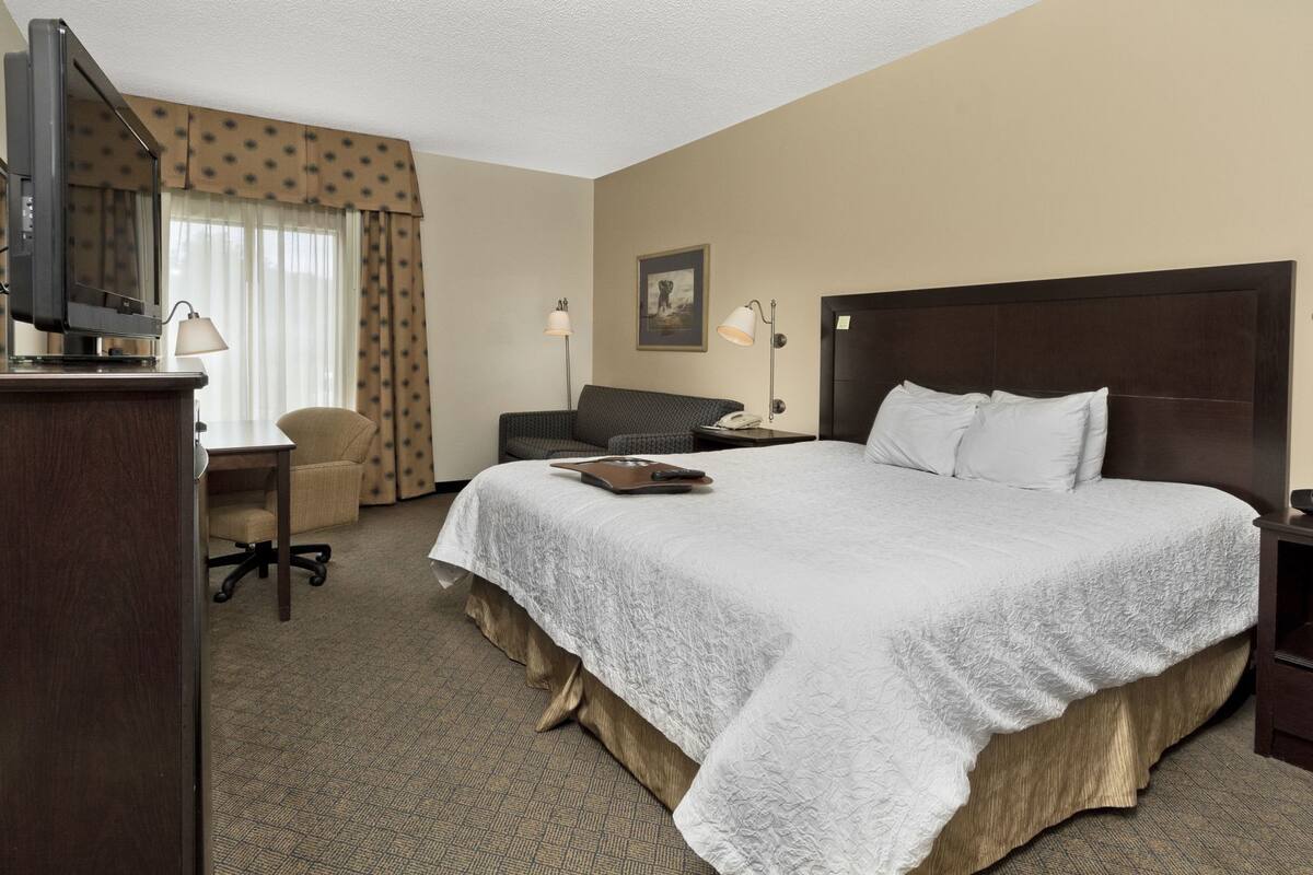 Ein Hotelzimmer mit einem großen Bett, einem Schreibtisch mit Computer, einem Sessel, einem Fernseher und einem Fenster mit Vorhängen.