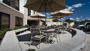 Un espace extérieur aménagé en terrasse, avec des tables et des chaises rondes, de grands parasols et un bâtiment en briques en arrière-plan.