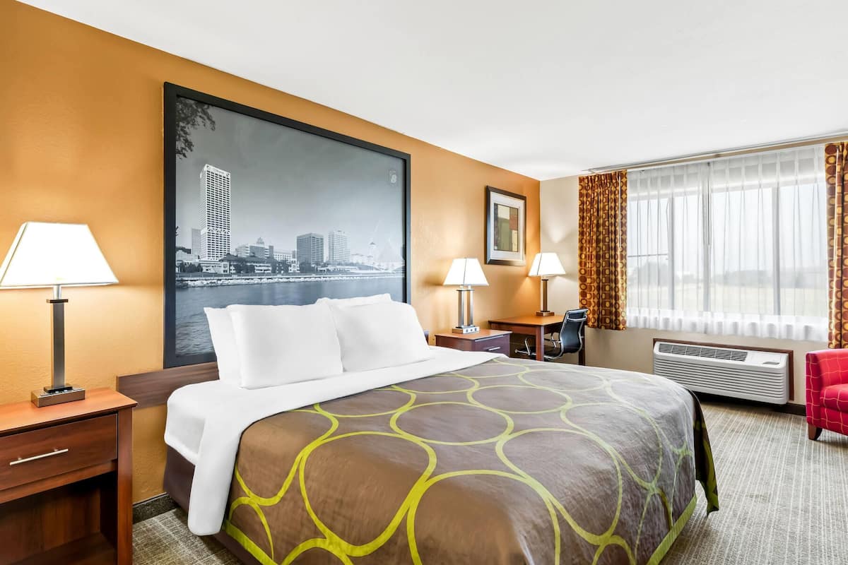 Une chambre d’hôtel avec un grand lit, deux tables de chevet, un bureau, une chaise, une fenêtre avec des rideaux et un tableau représentant un paysage urbain accroché au mur.