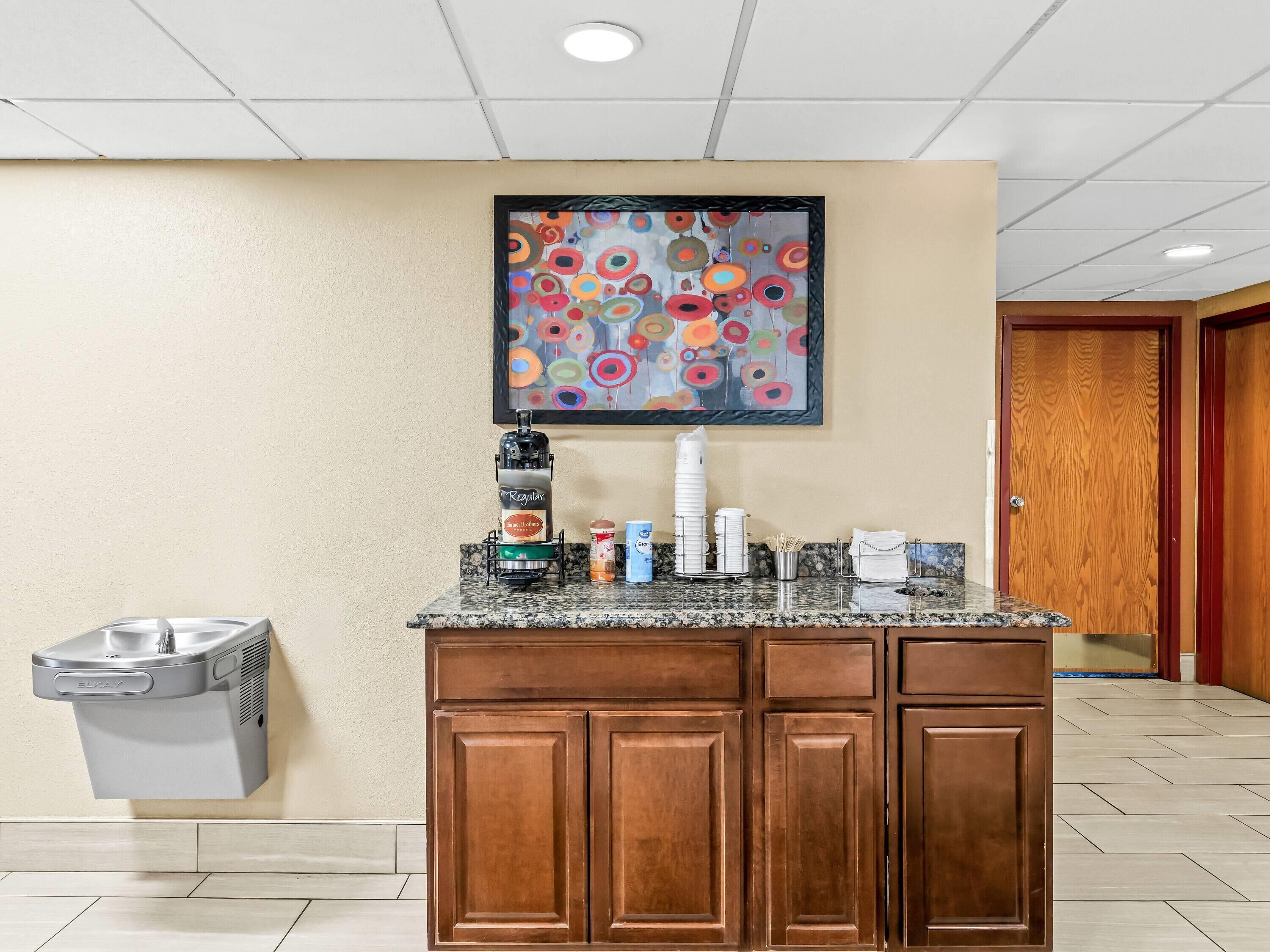 Una kitchenette de hotel con una cafetera, un cubo de basura y un cuadro en la pared.