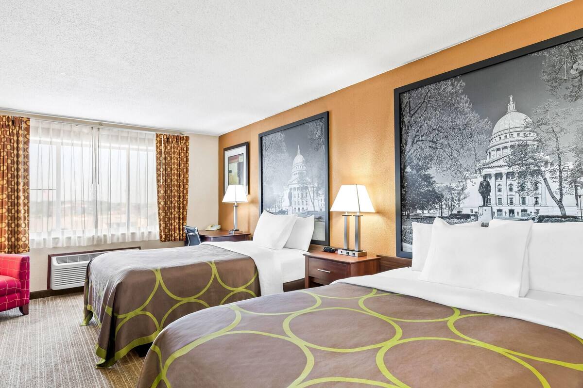 Une chambre d’hôtel avec deux lits, un bureau, une chaise, une lampe et un grand tableau encadré représentant un bâtiment.