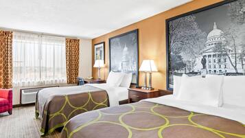 Une chambre d’hôtel avec deux lits, un bureau, une chaise, une lampe et un grand tableau encadré représentant un bâtiment.
