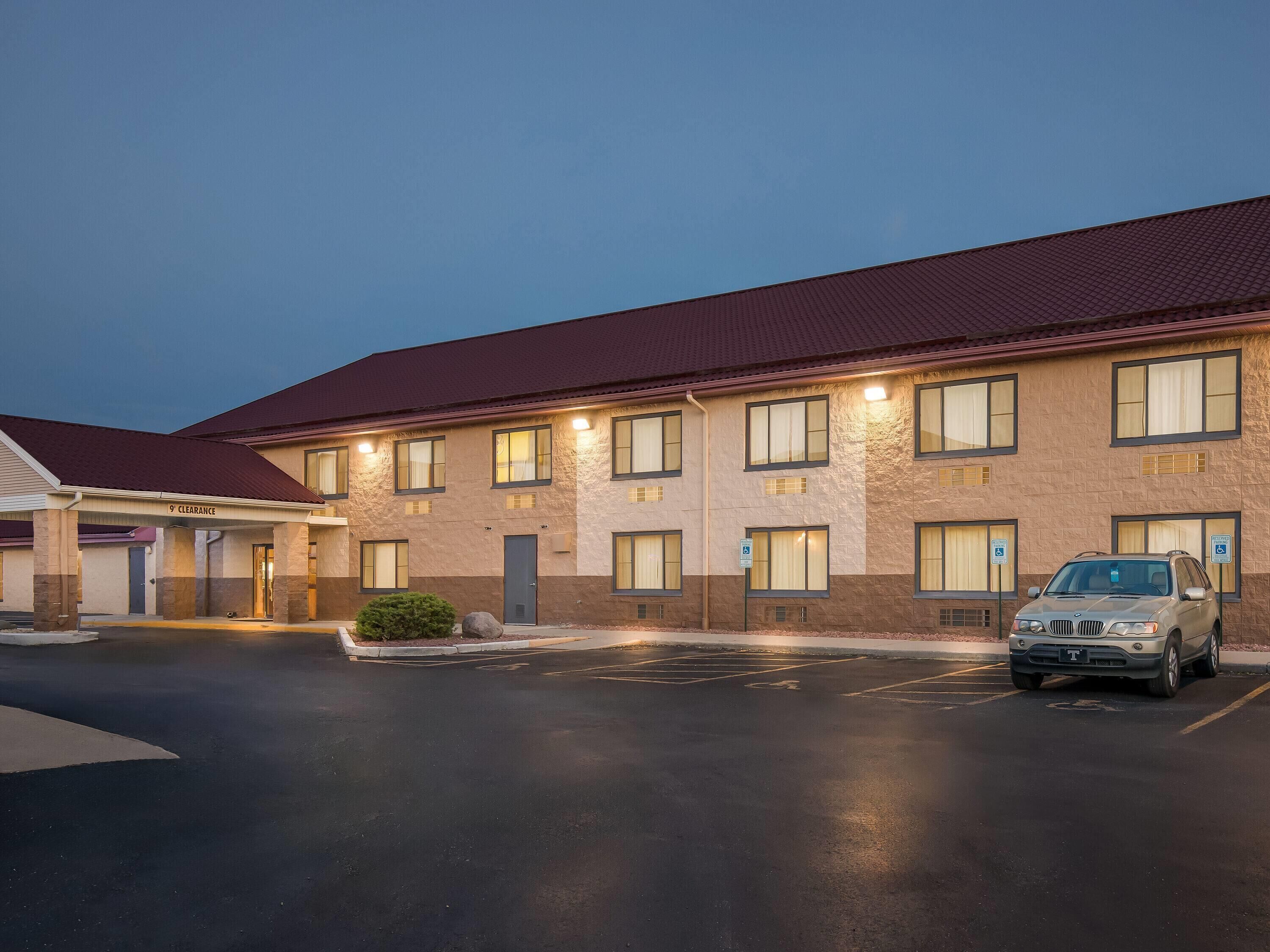 A multi-story hotel with a red roof and beige walls, featuring a covered entrance and a parking lot with a car.