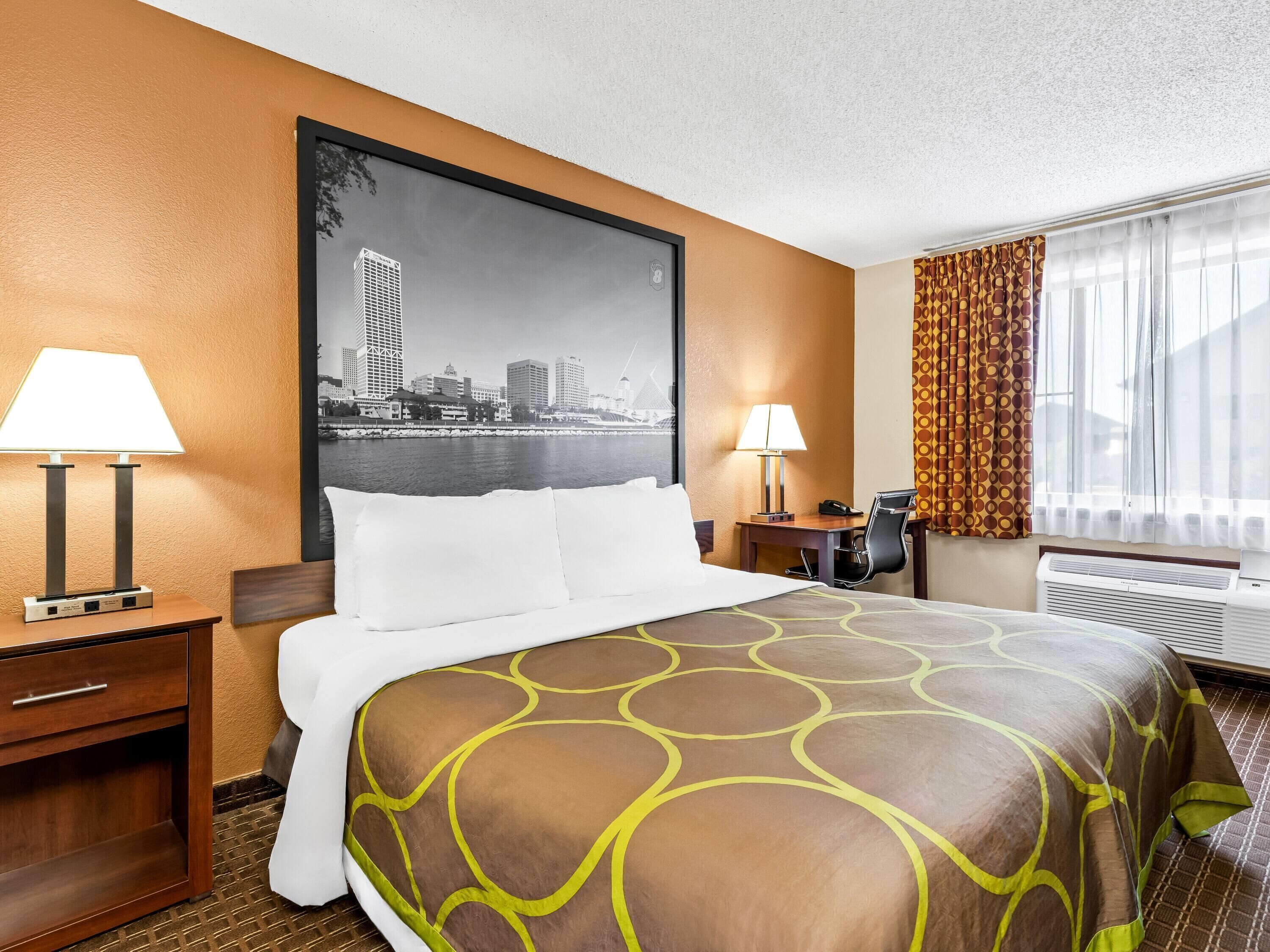 A hotel room with a bed, two bedside tables, a desk with a chair, a desk lamp, a telephone, and a large window with curtains.