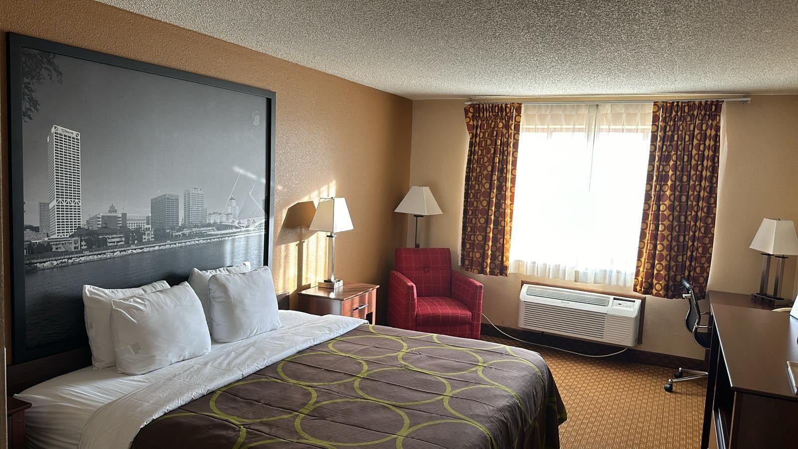 A hotel room with a large bed, a red armchair, a desk with a lamp, and a large window with curtains.